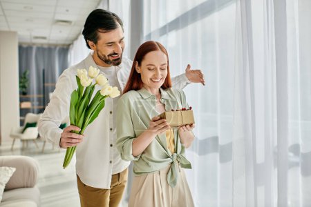Photo for A beautiful adult couple, a redhead woman and a bearded man, hold a gift box in front of a window in a modern apartment. - Royalty Free Image