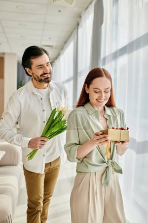 Téléchargez les photos : A bearded man and a redhead woman are walking through a modern living room, holding a gift box. - en image libre de droit