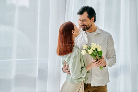 Photo for A redhead woman and bearded man holding flowers, basking in sunshine by a window in a modern apartment. - Royalty Free Image