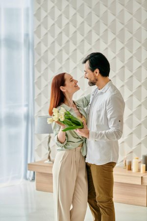 A redhead woman and bearded man stand in a room filled with flowers, enjoying quality time together in a contemporary setting.