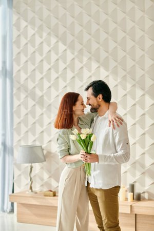 A redhead woman and bearded man stand in front of a wall with flowers in a modern apartment, enjoying quality time together.