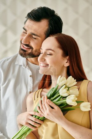 Photo for A redhead woman and bearded man, holding a bouquet of tulips, sharing a tender moment in a modern apartment. - Royalty Free Image