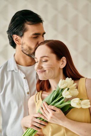 Foto de Una pelirroja y un hombre barbudo abrazan y besan mientras sostienen un ramo de tulipanes en un apartamento moderno. - Imagen libre de derechos
