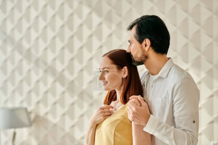 Foto de Una pareja de adultos con estilo, una mujer pelirroja y un hombre barbudo, de pie juntos frente a una pared blanca minimalista. - Imagen libre de derechos