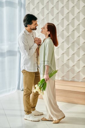 Photo for A redhead woman and bearded man embrace in a room adorned with colorful flowers, enjoying quality time together at home. - Royalty Free Image