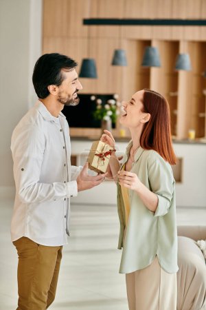 Photo for A beautiful adult couple, a redhead woman and bearded man, exchanging gifts in a modern apartment living room. - Royalty Free Image