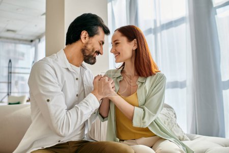 Photo for A beautiful adult couple, a redhead woman, and a bearded man, are sitting together on a couch in a modern apartment, enjoying quality time. - Royalty Free Image