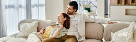 Photo for A beautiful adult couple, a redhead woman, and a bearded man, are sitting on a cozy couch in a modern living room, enjoying quality time together. - Royalty Free Image