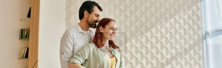 A stylish redhead woman and a bearded man stand together in front of a trendy wall in a modern apartment.