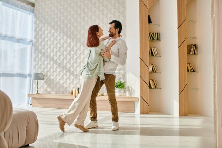 Foto de Una hermosa pelirroja y un hombre barbudo giran elegantemente y bailan en un moderno salón de apartamentos. - Imagen libre de derechos