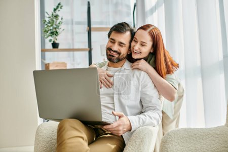 Photo for Adult couple, redhead woman and bearded man, enjoy quality time together while sitting on a couch and using a laptop. - Royalty Free Image