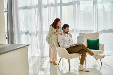 Foto de Una mujer pelirroja y un hombre barbudo se sientan en una silla, usando un portátil juntos en un apartamento moderno, disfrutando de un tiempo de calidad. - Imagen libre de derechos