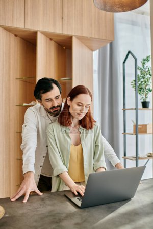 A beautiful adult couple, a redhead woman and a bearded man, working together on a laptop in a modern kitchen.
