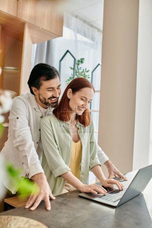 A redhead woman and bearded man engrossed in the screen of a laptop while standing in their modern kitchen.