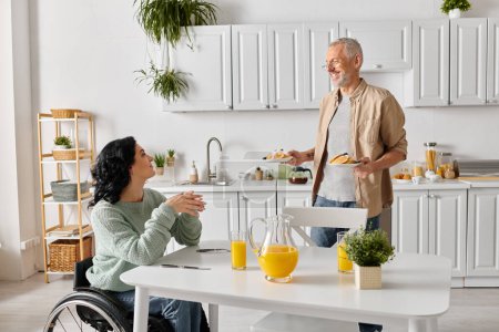 A disabled woman in a wheelchair engages in a conversation with her husband in a cozy kitchen setting at home.