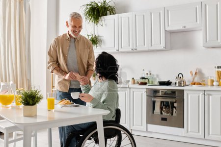 Foto de Un marido está junto a su esposa discapacitada en una silla de ruedas en la acogedora cocina de su casa, compartiendo un momento de convivencia. - Imagen libre de derechos