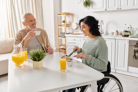 A disabled woman in a wheelchair and her husband share a warm moment at the kitchen table in their home.