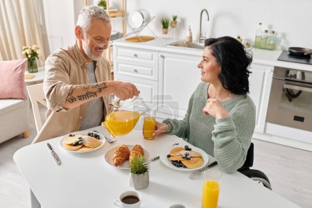 A disabled woman in a wheelchair and her husband enjoy breakfast together at a kitchen table in their cozy home.
