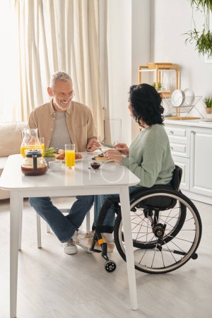 A man in a wheelchair and a woman at a table in their kitchen at home, sharing a moment of togetherness.
