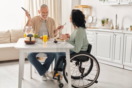 Photo for A man in a wheelchair and a woman enjoy a meal together in their kitchen at home. - Royalty Free Image