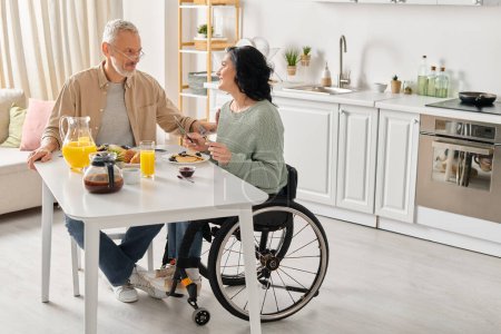 Téléchargez les photos : Un homme en fauteuil roulant et une femme assis ensemble à une table dans leur cuisine à la maison. - en image libre de droit