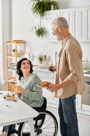 A woman in a wheelchair happily looking at husband holds a plate with croissant in a cozy kitchen setting at home.