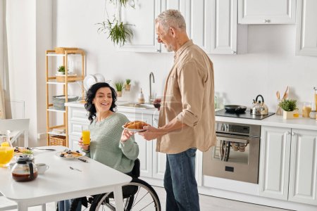 Foto de Una mujer en silla de ruedas sosteniendo un plato de croissant mientras interactúa con su pareja en una acogedora cocina en casa. - Imagen libre de derechos