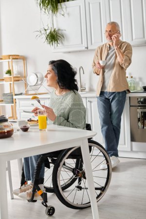 Téléchargez les photos : Un mari et sa femme handicapée, en fauteuil roulant, se tiennent côte à côte à une table de cuisine - en image libre de droit