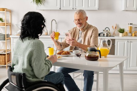 Una mujer en silla de ruedas y un hombre se sientan juntos en una mesa en la cocina de casa, disfrutando de una comida y de la compañía de los demás..