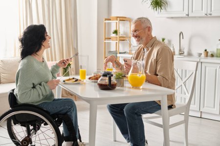 Téléchargez les photos : Une femme handicapée en fauteuil roulant et son mari s'assoient à une table, prenant le petit déjeuner dans leur cuisine à la maison. - en image libre de droit