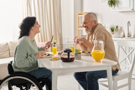 A man and woman in wheelchair engaged in conversation in a domestic kitchen setting.