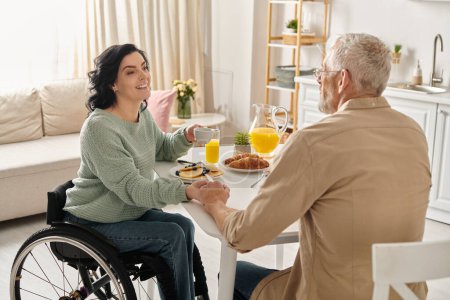 Foto de Una mujer discapacitada en silla de ruedas está siendo servida cariñosamente con jugo de naranja por su marido en su cocina casera. - Imagen libre de derechos