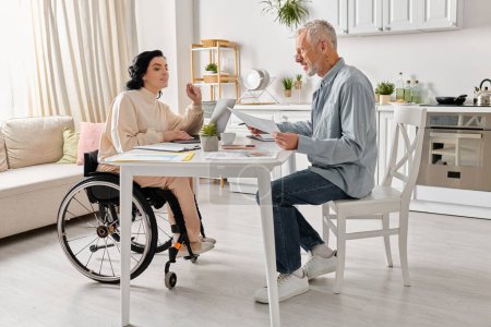 Téléchargez les photos : Un homme assis à une table à côté d'une femme en fauteuil roulant dans leur cuisine à la maison. - en image libre de droit