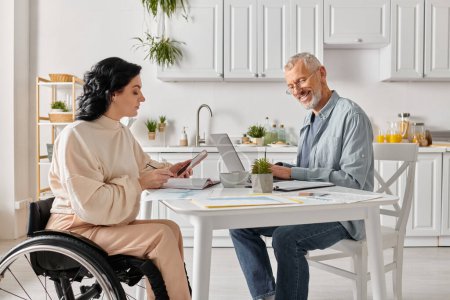 Téléchargez les photos : Une femme en fauteuil roulant et son mari utilisant un ordinateur portable dans la cuisine à la maison. - en image libre de droit