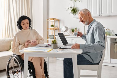 Une femme en fauteuil roulant et un homme avec un ordinateur portable, partageant un moment de convivialité dans leur cuisine à la maison.