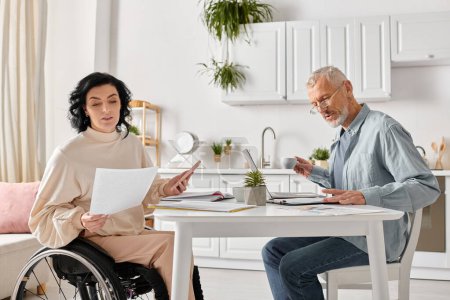 Une femme dans un fauteuil roulant tenant le téléphone et un homme avec un ordinateur portable, partageant un moment de convivialité dans leur cuisine à la maison.