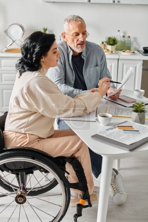 Une femme handicapée en fauteuil roulant et son mari confortablement assis à une table dans leur cuisine à la maison.