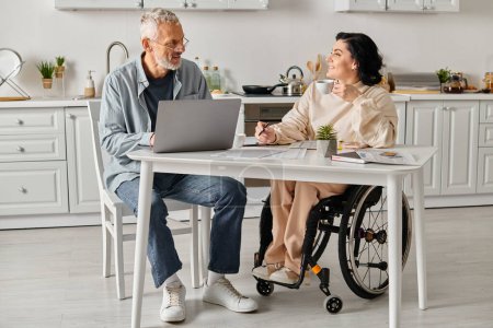 Téléchargez les photos : Un mari et sa femme handicapée sont assis à une table de cuisine travaillant ensemble sur un ordinateur portable. - en image libre de droit
