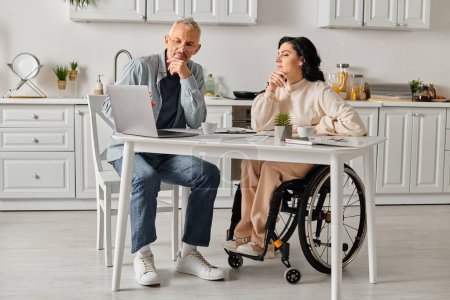 A woman in a wheelchair and her husband sit at a table, engaged with a laptop in a cozy kitchen setting at home.