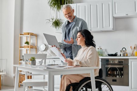 A man and woman in a wheelchair engrossed in a laptop screen in a cozy kitchen at home.