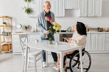 A man lovingly hands flowers to a woman in a wheelchair, surrounded by a cozy kitchen at home.