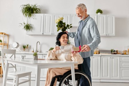 A man standing devotedly next to his disabled wife in a wheelchair in their kitchen at home.