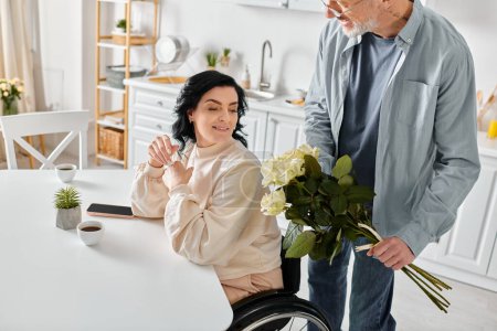 Téléchargez les photos : Un homme tenant un bouquet, se tient amoureusement à côté de sa femme alors qu'elle est assise dans un fauteuil roulant dans leur cuisine à la maison. - en image libre de droit