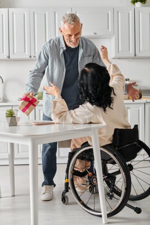 A man gives a gift to his wife in a wheelchair, in the kitchen of their home.