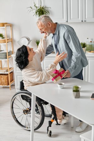 Photo for A disabled woman in a wheelchair touching her husbands hand lovingly in the kitchen at home, receiving present - Royalty Free Image