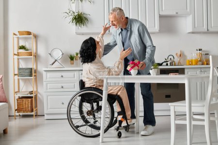 A loving man gives a gift to his happy wife in a wheelchair, in the kitchen of their home.