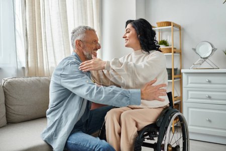 Téléchargez les photos : Une femme en fauteuil roulant étreint son mari, montrant de l'amour et du soutien dans leur salon. - en image libre de droit