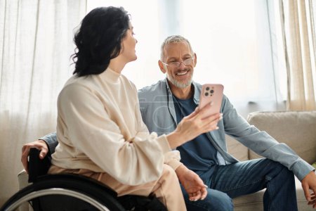 Téléchargez les photos : Une femme handicapée en fauteuil roulant tient un téléphone cellulaire alors qu'elle est assise dans son salon, son mari à ses côtés. - en image libre de droit