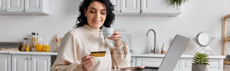 Una mujer feliz tiene una tarjeta de crédito mientras se centra en su computadora portátil en la cocina.