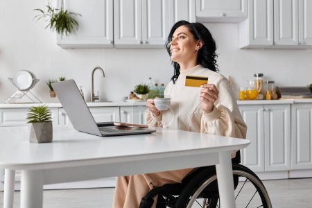 A woman in a wheelchair confidently shops online with a credit card and laptop in her kitchen.